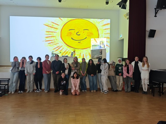 Die Autorin Birgit Gröger, der Autor Daniel Schneider, die Lehrkraft Marielle Niebisch und die Schülerinnen und Schüler des WPU Kurses stehen in der Aula vor der Bühne. Über ihnen ist auf der Leinwand eine große Sonne zu sehen. 