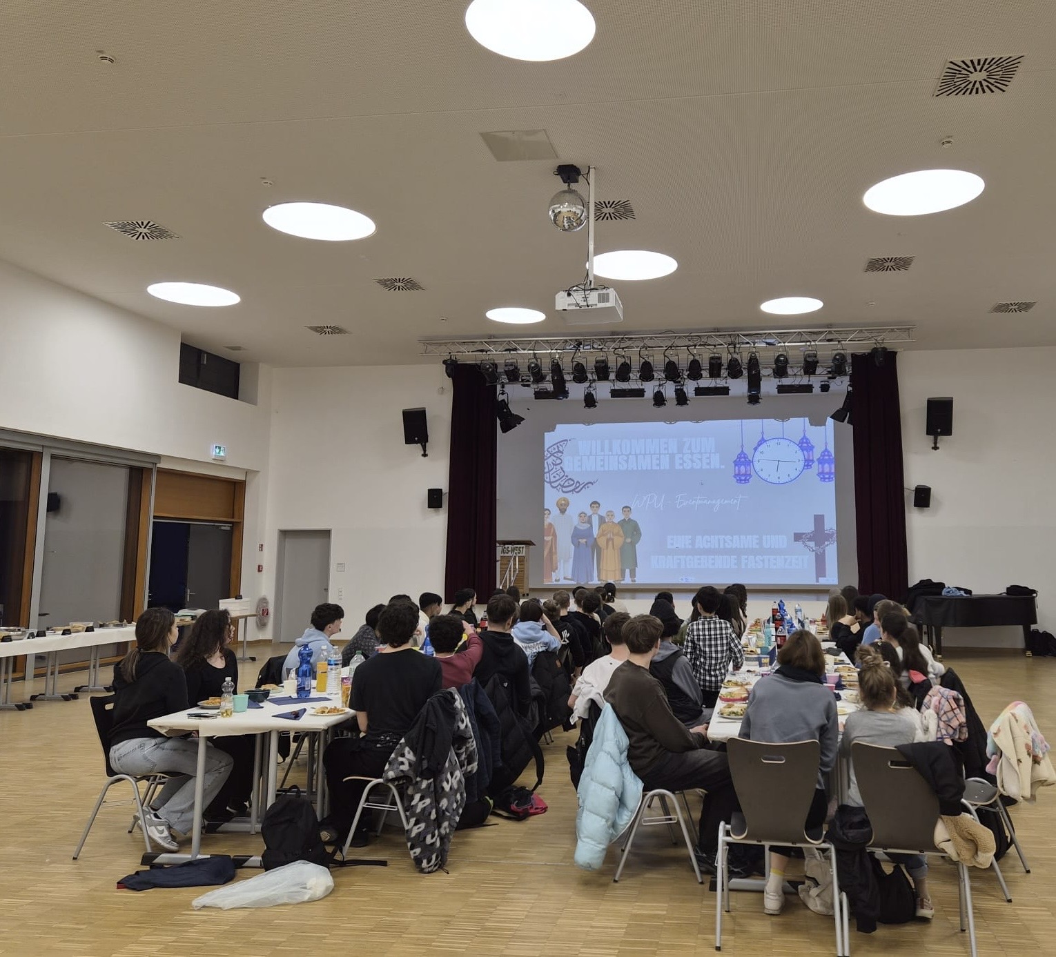 Schülerinnen und Schüler der Jahrgangsstufe 10 und ihre Lehrkräfte essen und trinken zusammen in der Aula