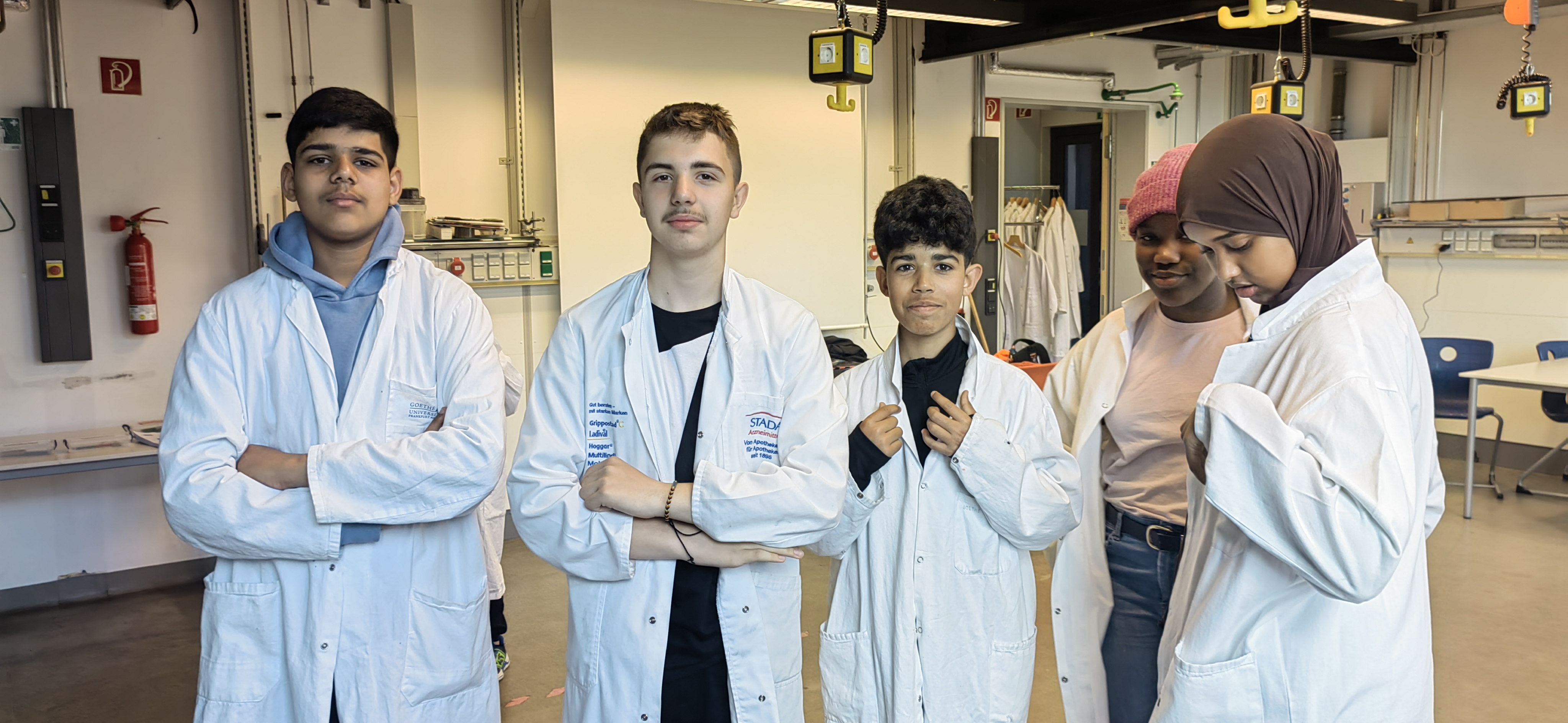 Schülerinnen und Schüler der Intensivklasse stehen im Labor der Goethe-Universität. Sie tragen Labor-Kittel. 