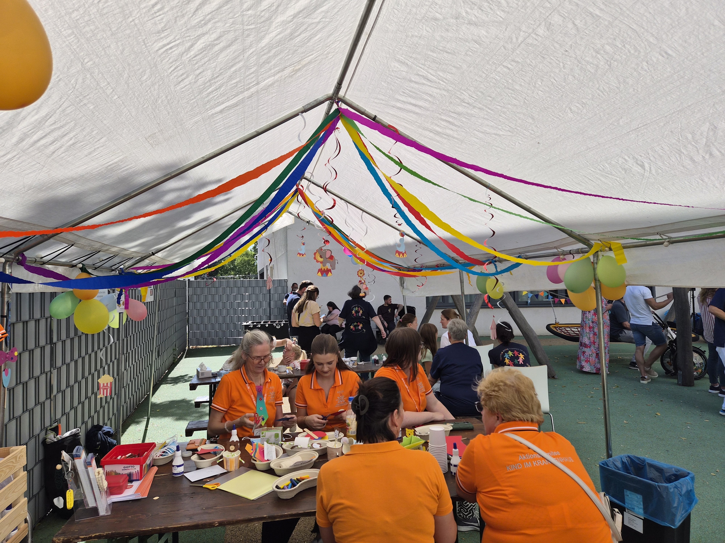 mehrere ehrenamtliche Personen, die unterm Zelt am Basteltisch mit Kindern kreativ arbeiten